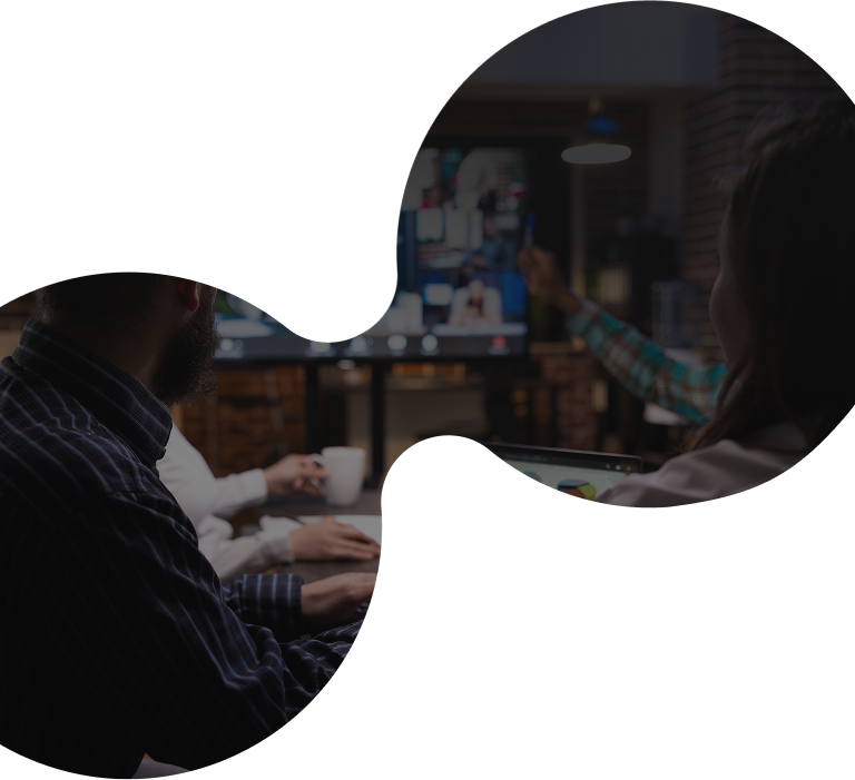 Two people sit at a desk, viewing and discussing information on a large screen in a dimly lit office. The image is partially obscured by a white, abstract, curved shape overlay.