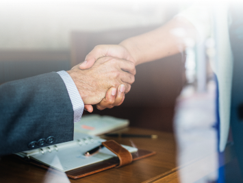 Two people shaking hands across a desk, one wearing a suit. An open planner, pen, and other office items are visible on the desk, suggesting a business meeting or agreement.