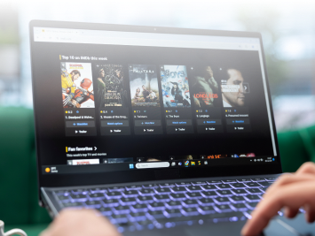 A person using a laptop with a movie streaming website open, displaying various film posters and titles on the screen. The keyboard is illuminated, and the users hands are partially visible.