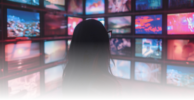 A person with long hair and glasses is seen from behind, looking at a wall of multiple colorful screens displaying various images in a dimly lit room.