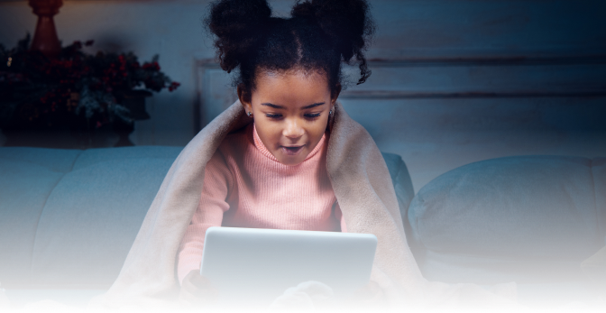 A young girl with curly hair sits on a couch under a blanket, looking at a tablet screen with a focused and engaged expression.