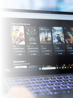 A laptop screen displays a streaming website showing movie thumbnails, ratings, and titles in a row, with a blurry keyboard visible at the bottom of the image.